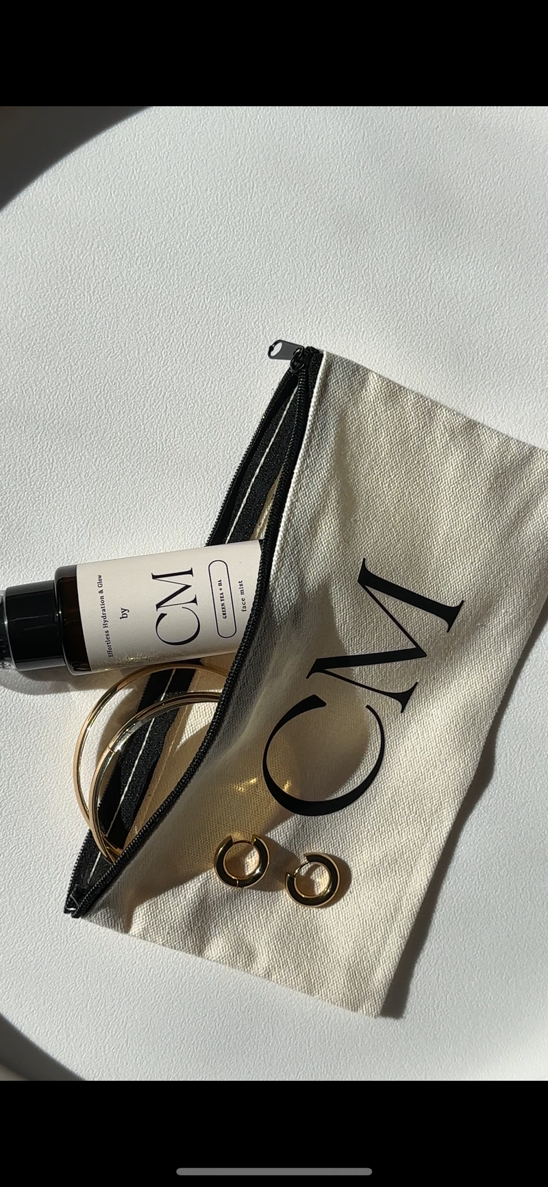 Cosmetic bag with 'CM' branding, a bottle, and makeup items on a white surface.